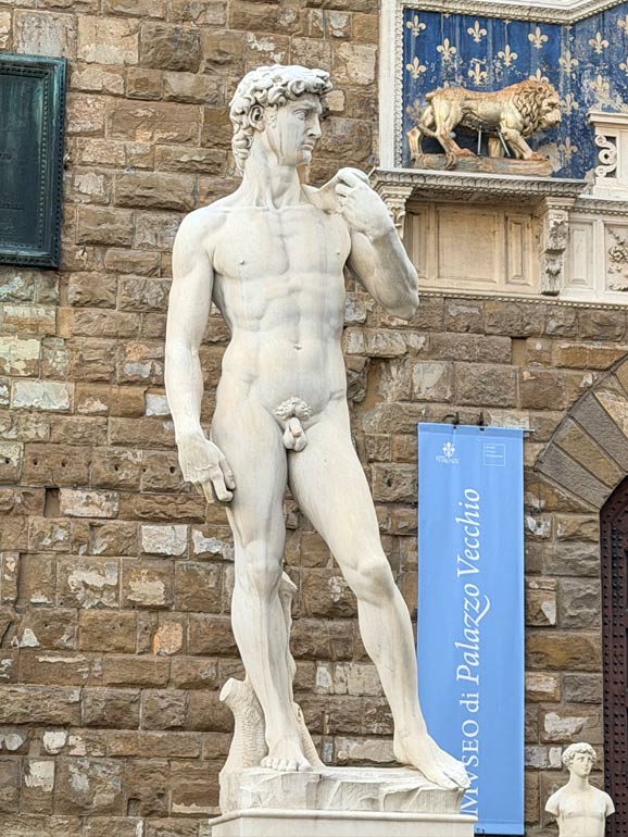Statue of David. White in color, nude male figure. Outside in front of building.