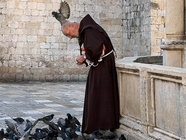 A friar feeding pigeons with a pigeon on his head.