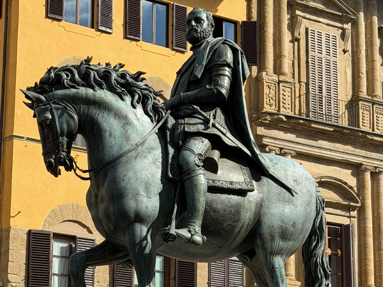 Monument of Cosimo 1 statue. Green in color with man with beard riding a horse. Yellow building in background.