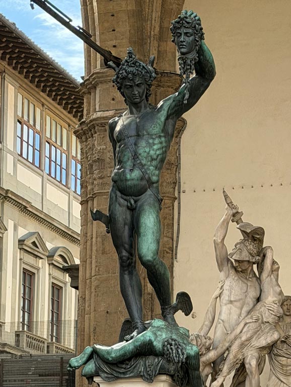 Green statue with man holding up woman's head. Her body is between his legs. Outside in front of building.