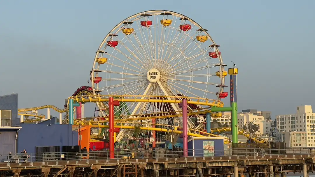 Santa Monica Pacific Wheel glowing with multicolored LED lights on the Santa Monica Pier at sunset, overlooking the Pacific Ocean with waves below and a pastel sky in the background.
