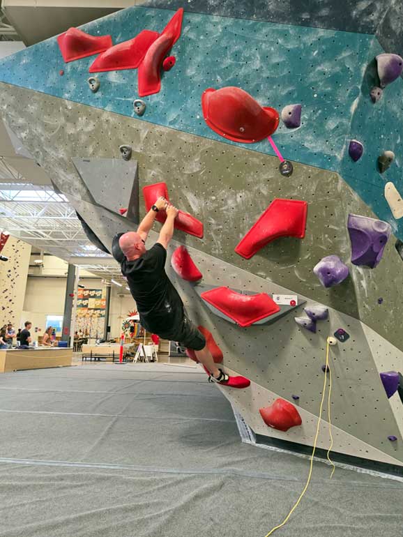 Gordon Climbing Third Route Gordon Eaton IV coiled and generating power on an overhanging section of the wall, looking upward intently as he prepares to make a dynamic jump move toward a distant red hold during the Mile High Melee finals.