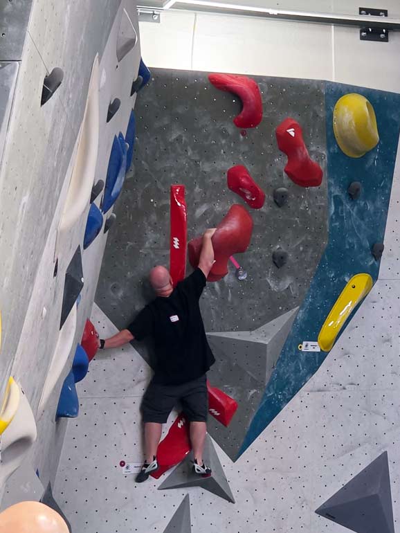 Gordon climbing the second Comp Route Gordon Eaton IV climber in athletic gear navigating a complex sequence of red climbing holds on a textured gym wall, finding his footing while competing in the Mile High Melee bouldering competition.