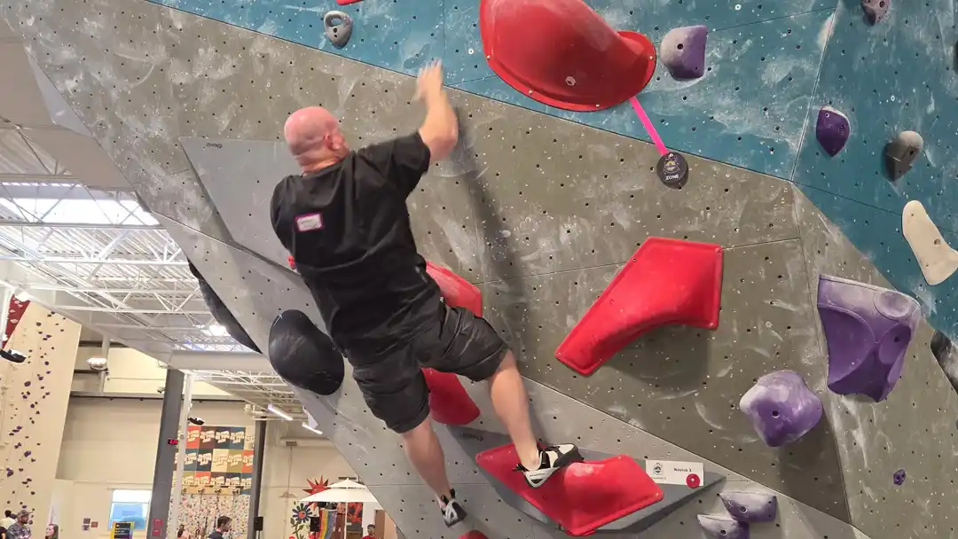 A dynamic action shot of a 51-year-old male climber launching himself mid-air toward a distant hold on a steep, overhanging bouldering wall. He is fully extended, reaching with fingertips extended toward a red plastic climbing grip during the Mile High Melee finals at Movement Centennial.