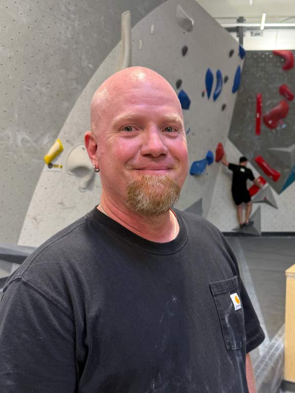 gordon-eaton-iv-mile-high-melee-finals-movement-centennial A portrait of Gordon Eaton IV smiling at the Mile High Melee bouldering competition finals inside Movement Centennial climbing gym, wearing climbing gear with the brightly lit, hold-covered bouldering walls blurred in the background.